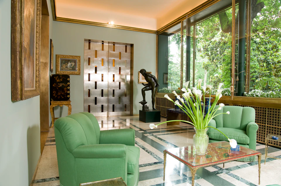 Room with green armchairs, glass coffee table with white flowers, large windows, a bronze statue, and a metallic accent wall.