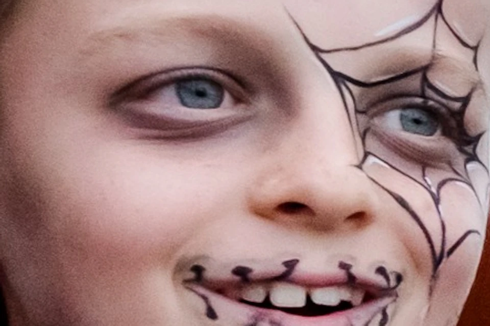 Child with face paint sits securely on a roller coaster ride, smiling, with a wooden amusement park visible in the background.