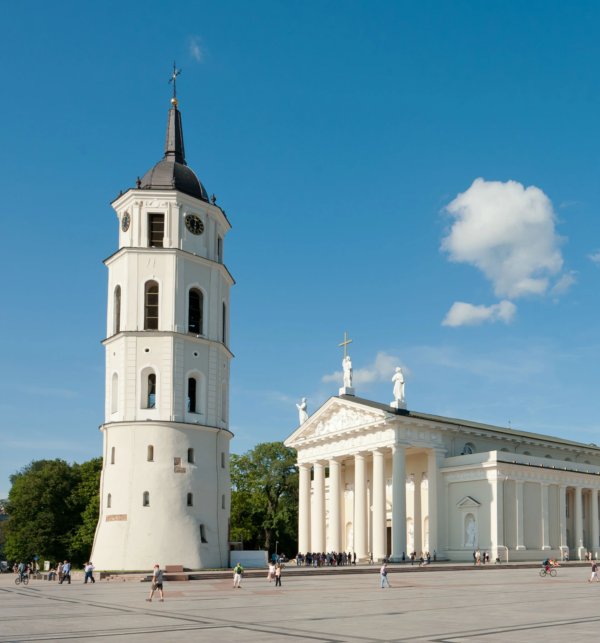 A historic tower and a white neoclassical building with columns under a clear blue sky, people walking in the plaza below.