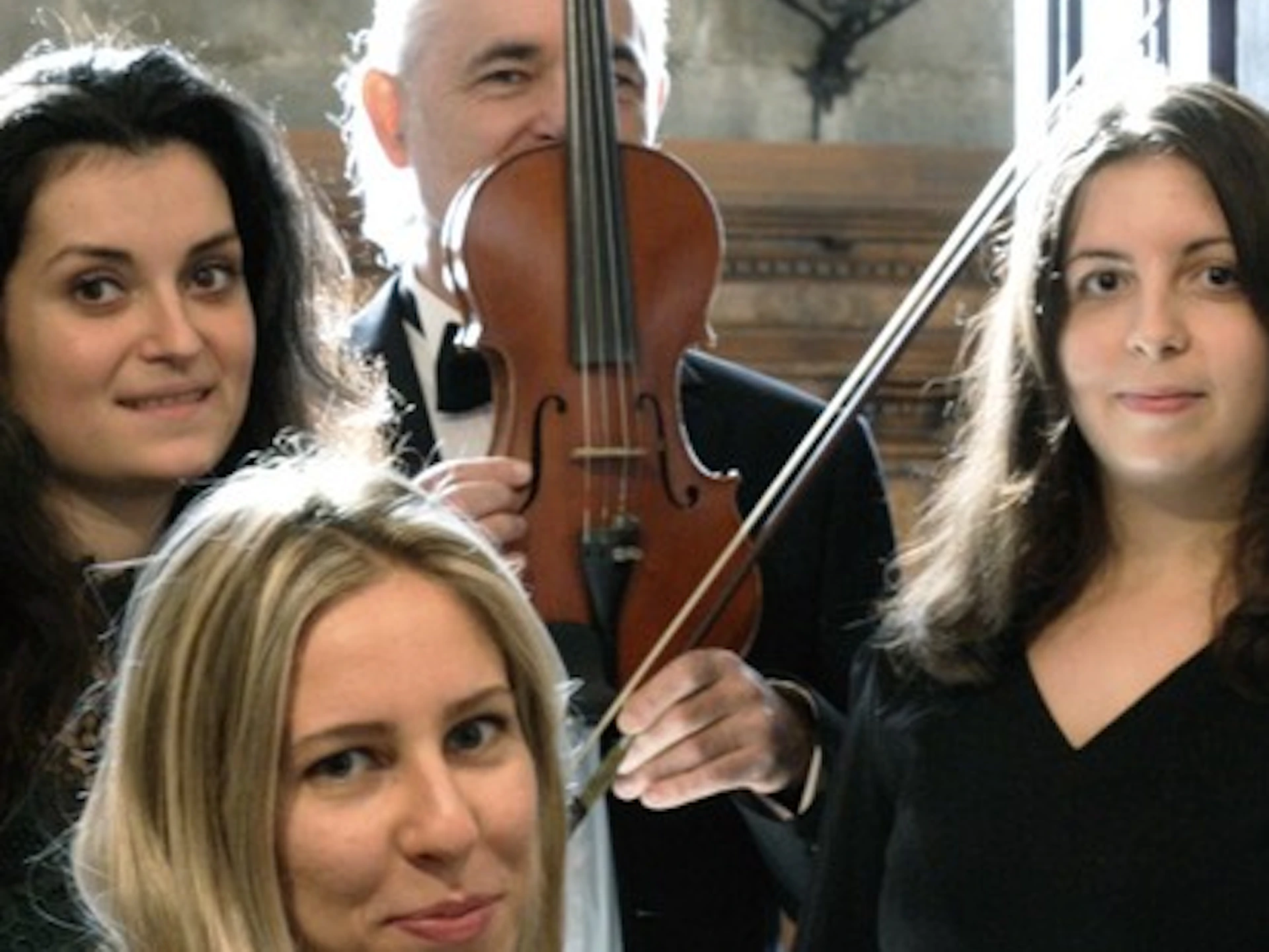 Four musicians with string instruments and a piano pose in a room with a candelabra and wooden decor.