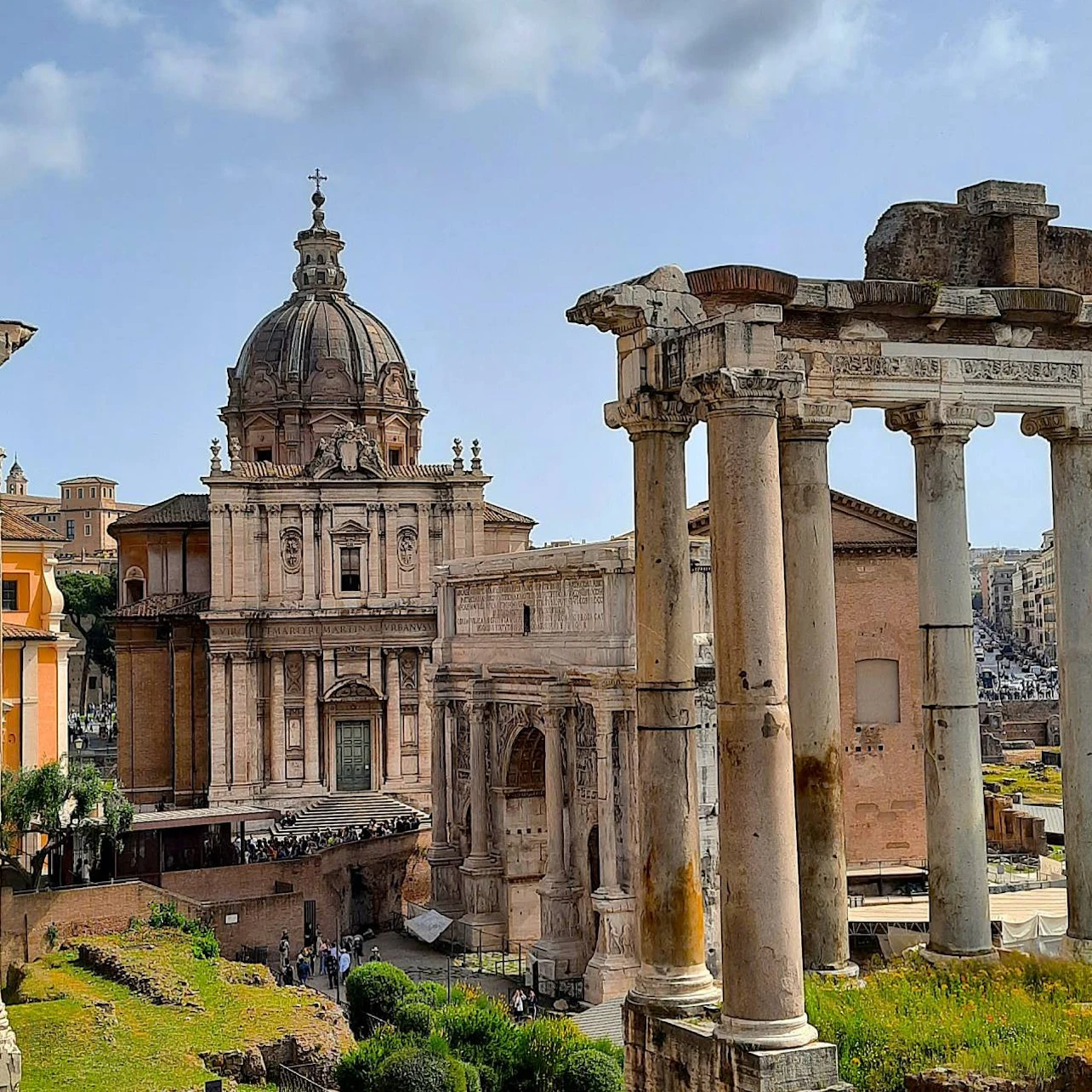Ancient Roman ruins with columns in the foreground and a historic domed building in the background under a partly cloudy sky.