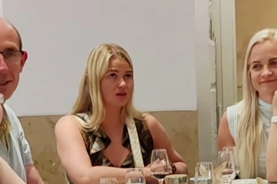 A group of people seated at a table indoors, with wine glasses and snacks. One holds a sign reading 'Lisbon Foodie Walk.'