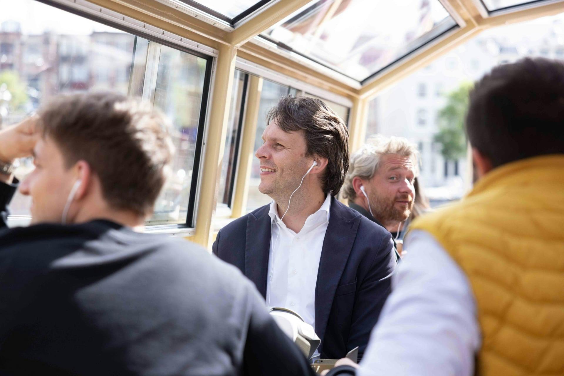 Drei Personen sitzen in einem Boot oder einer Straßenbahn mit Glasdach; zwei Männer tragen Kopfhörer, einer im Anzug, der andere in Freizeitkleidung.