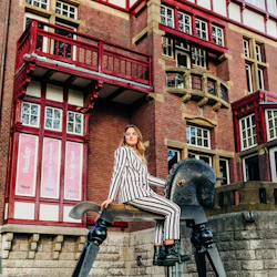 En person i randig kostym sitter på en stor gunghästskulptur, med en röd tegelbyggnad i bakgrunden.