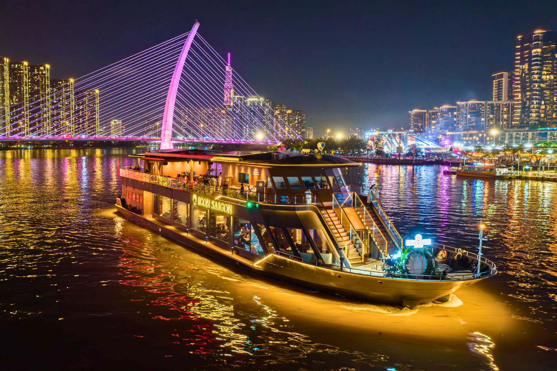 A lit-up cruise boat named Moon Saigon on a river at night, with a cable-stayed bridge and city lights in the background.