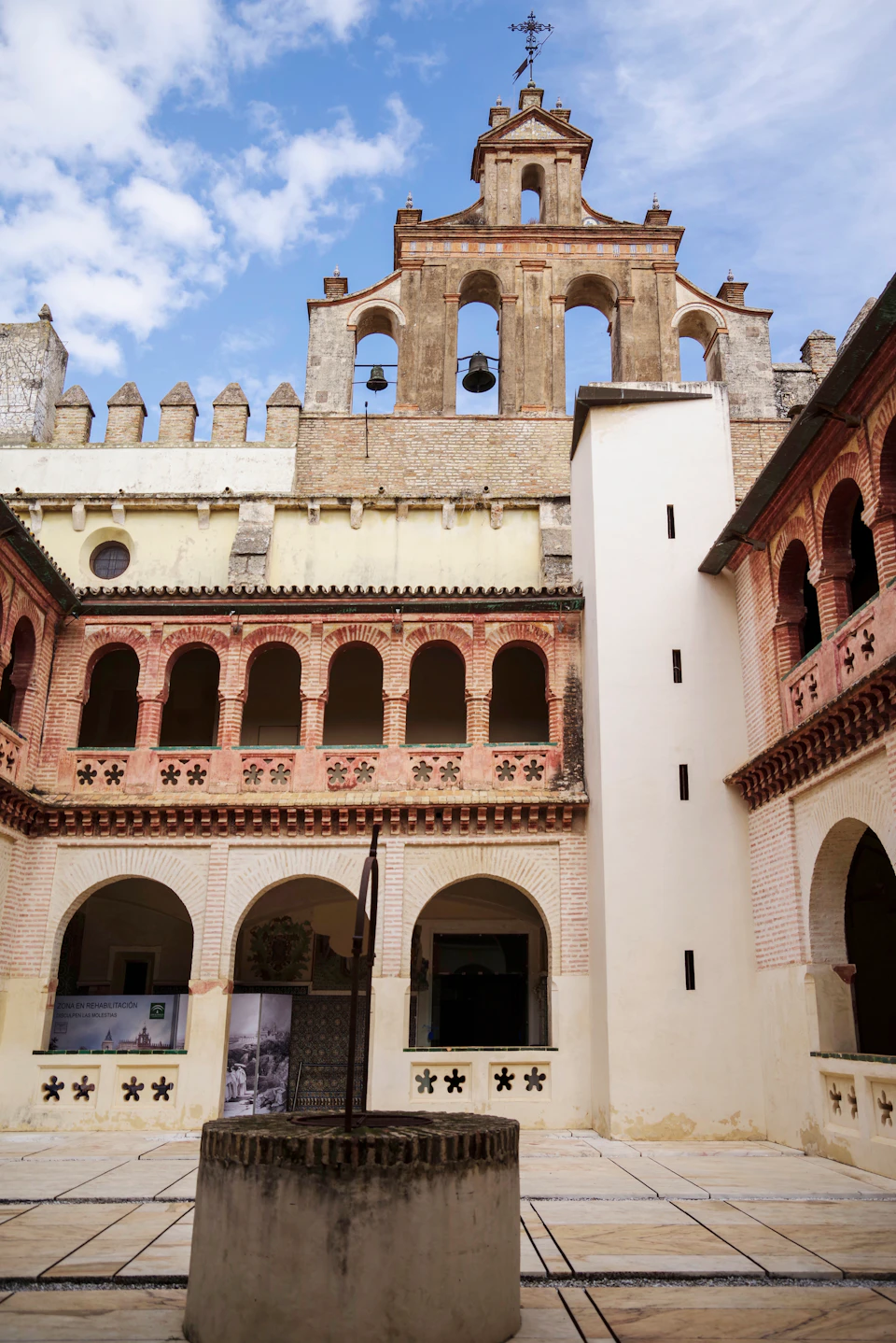 A historic courtyard with arched windows, brick walls, and a bell tower above under a partly cloudy sky.