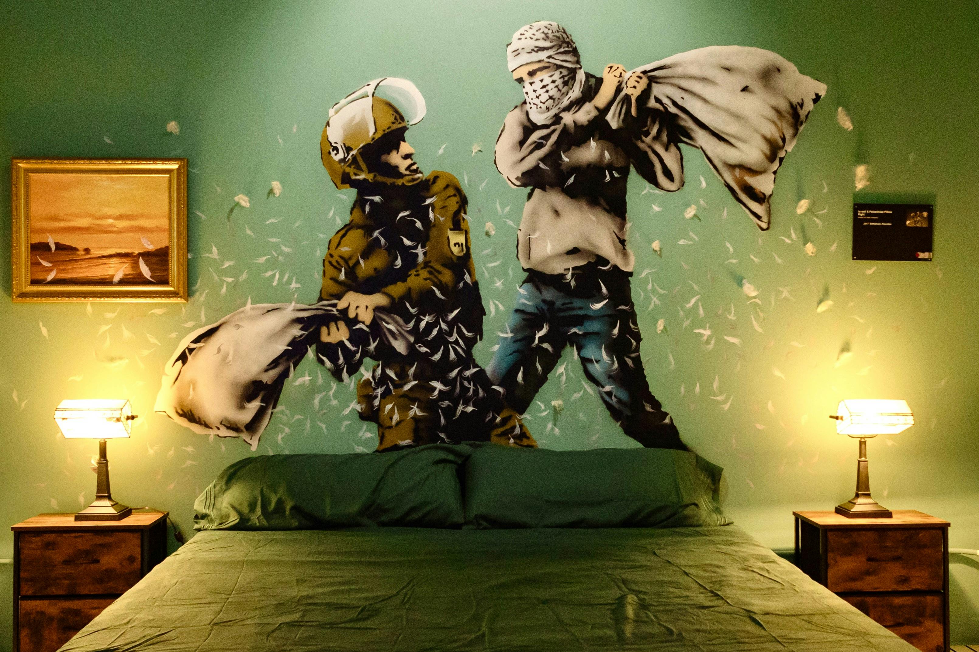 A bedroom with a green bedspread features a mural of a pillow fight between a soldier and a masked civilian on the wall.