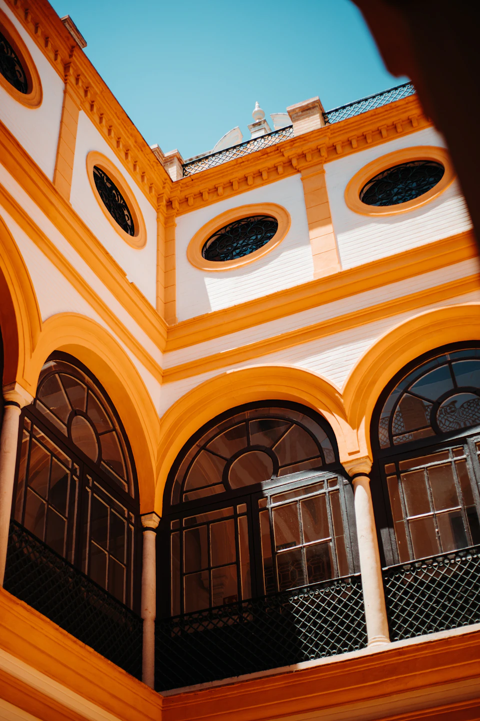 Inner courtyard with yellow arches, large rounded windows with black frames, and circular upper windows against a blue sky.