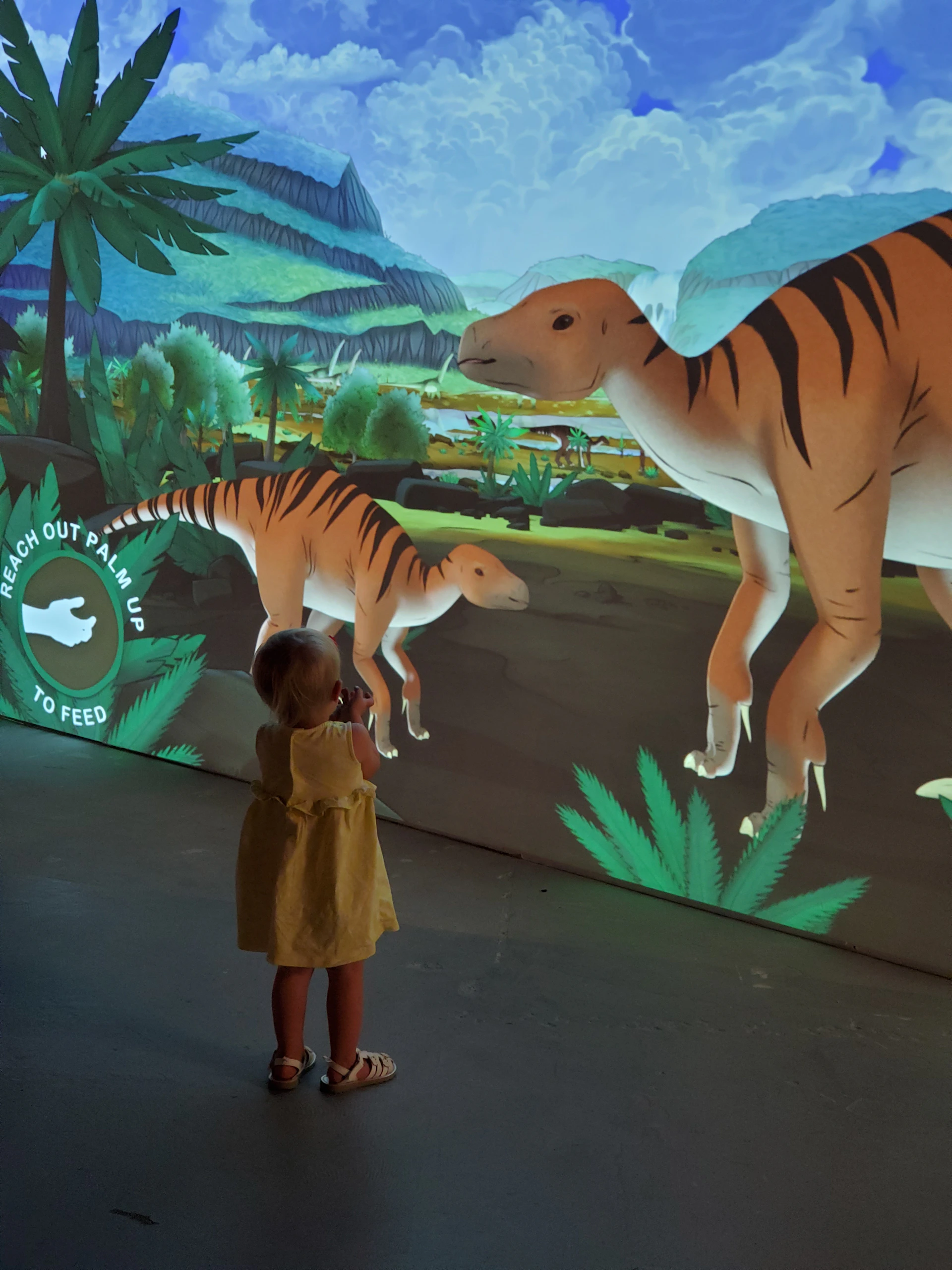 A child in a yellow dress interacts with a digital display of dinosaurs in a lush prehistoric landscape. Text on display reads, "Reach out palm up to feed."