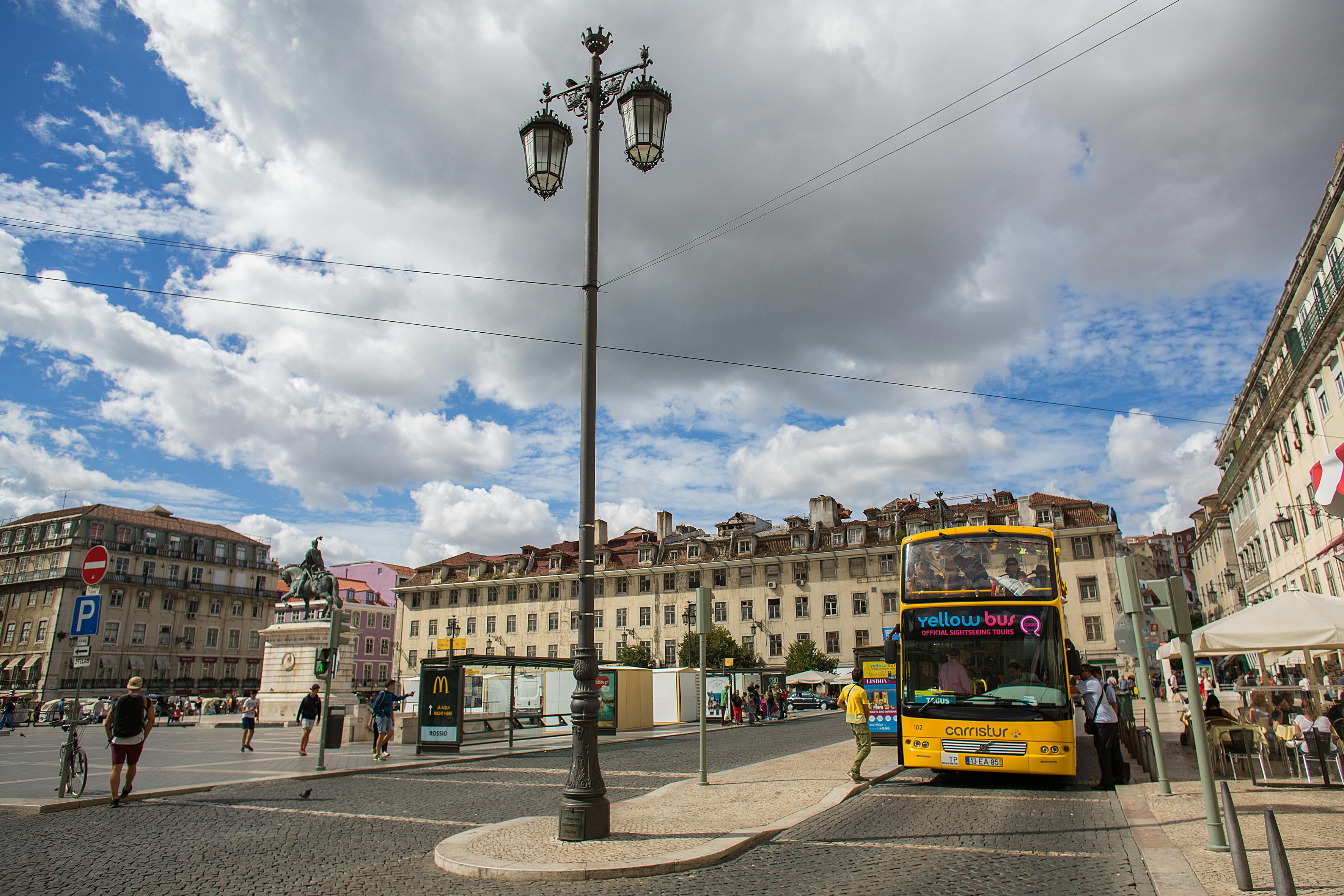 Brukowany plac z posągiem, starymi budynkami, lampą uliczną, chodzącymi ludźmi i żółtym autobusem piętrowym.