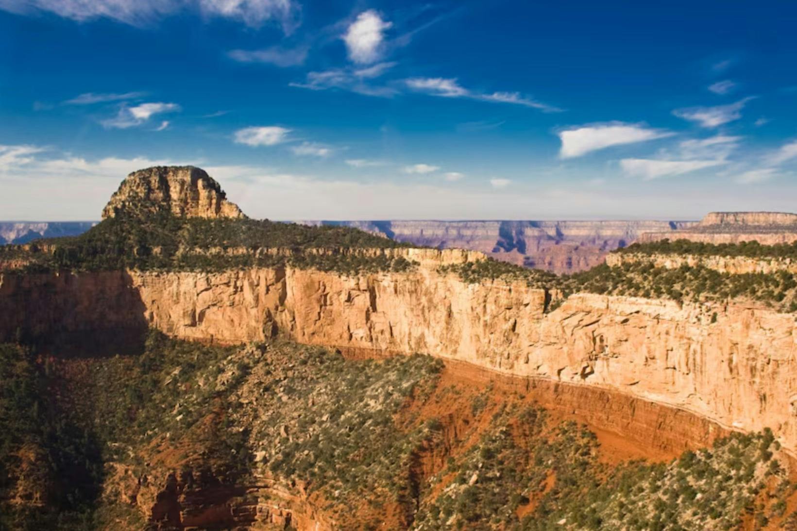 A scenic view of a desert canyon with steep rocky cliffs, sparse vegetation, and a bright blue sky with scattered clouds.