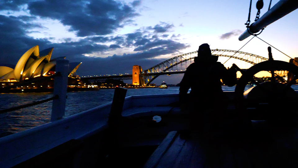 Silhouette of a person on a boat in front of the Sydney Harbour Bridge at dusk with city lights and cloudy sky in the background.