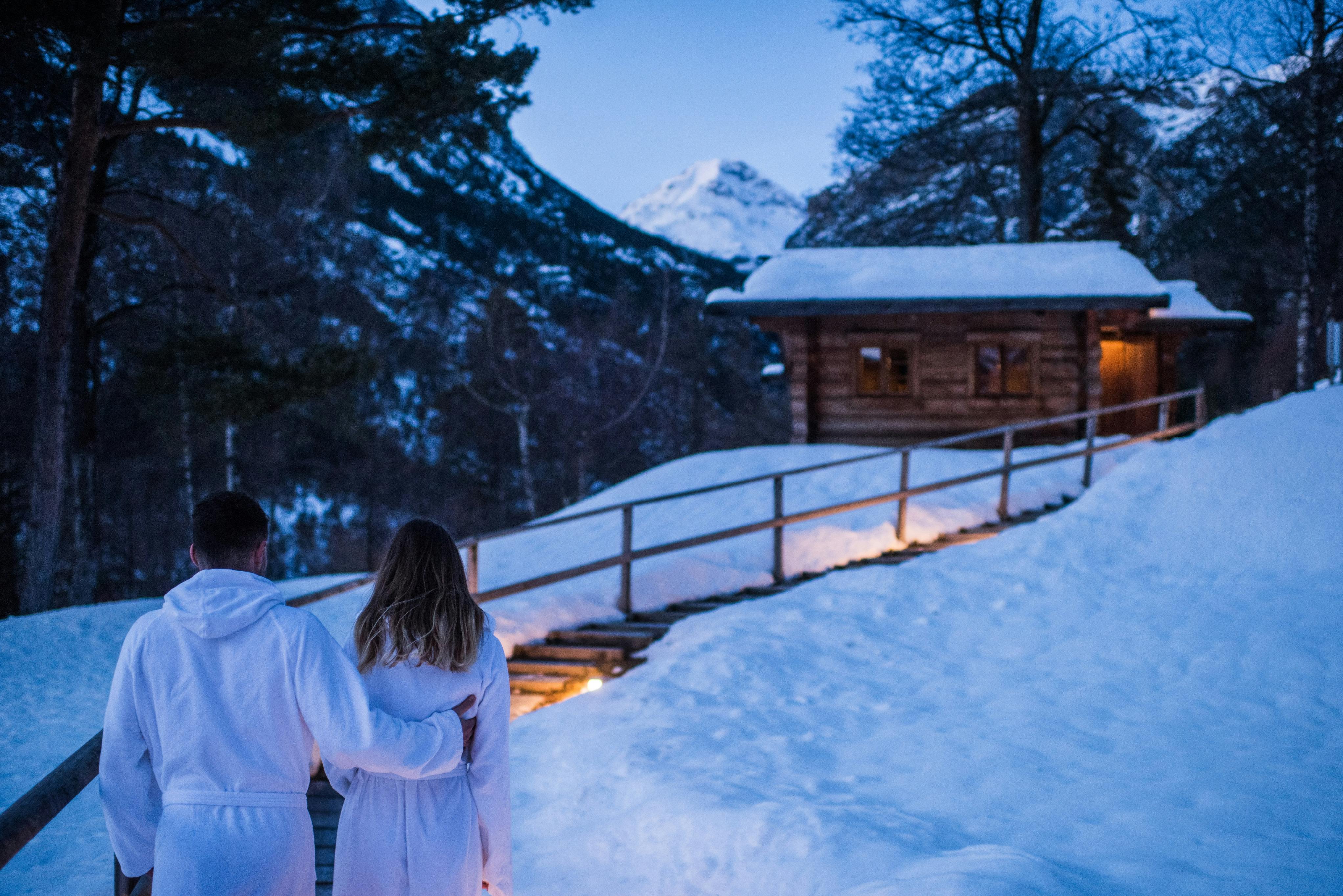 Una coppia in abiti bianchi che cammina verso una baita di legno illuminata in una foresta montuosa e innevata al tramonto.