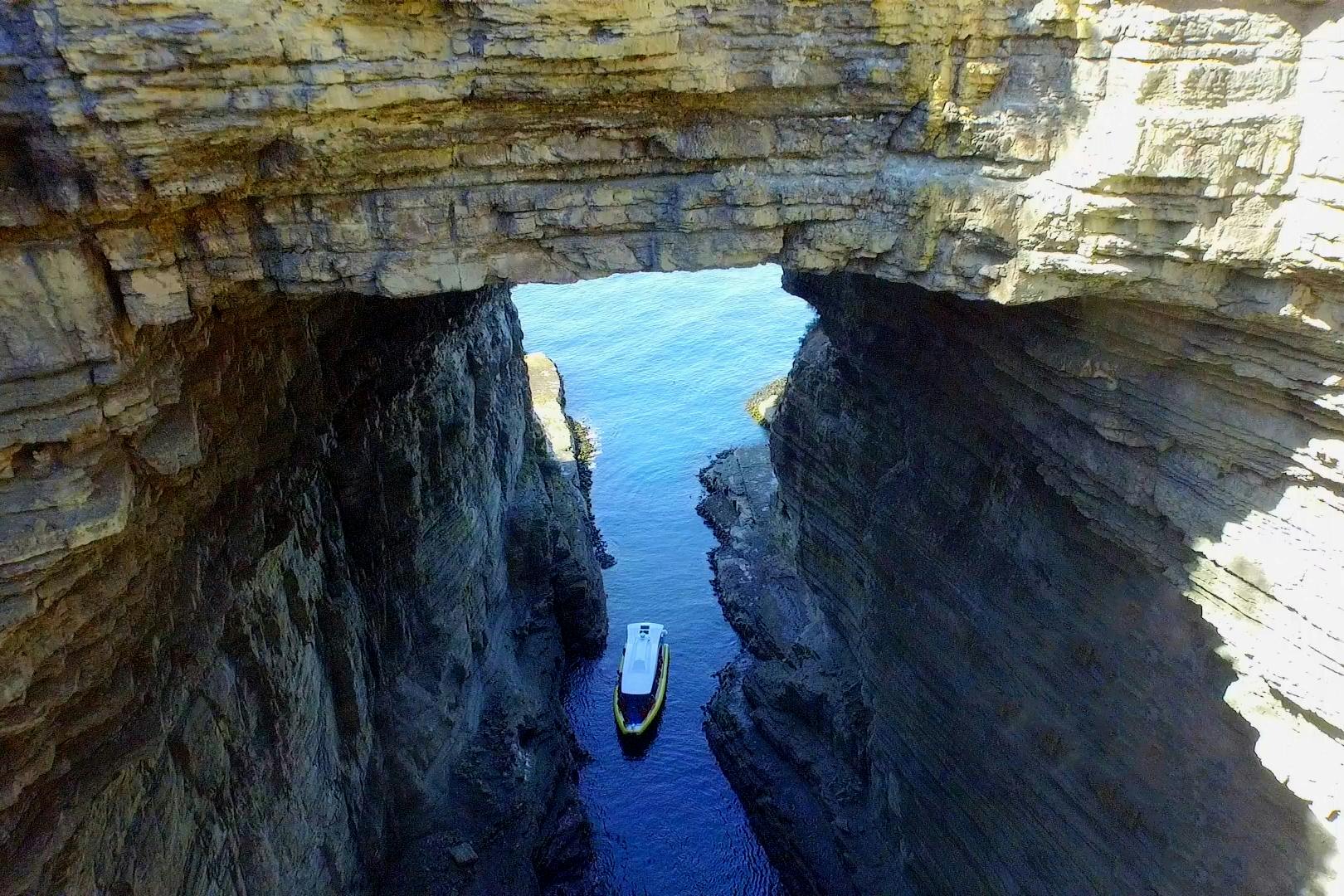 A small boat navigates through a narrow water passage between high rock cliffs under a natural rock bridge.