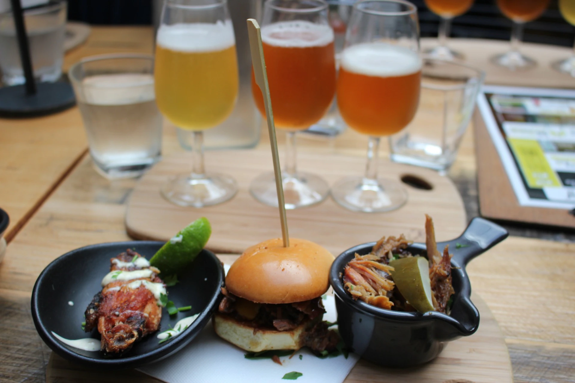 A meal with a small sandwich, meat dishes, and a flight of beer served on a wooden table.