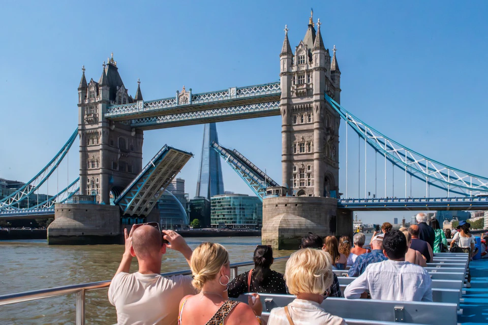 Tourists on a boat view Tower Bridge in London, opening to let a vessel through, with modern buildings in the background.