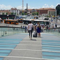 Two people are walking down a glass bridge toward a bus terminal with several buses and buildings in the background.