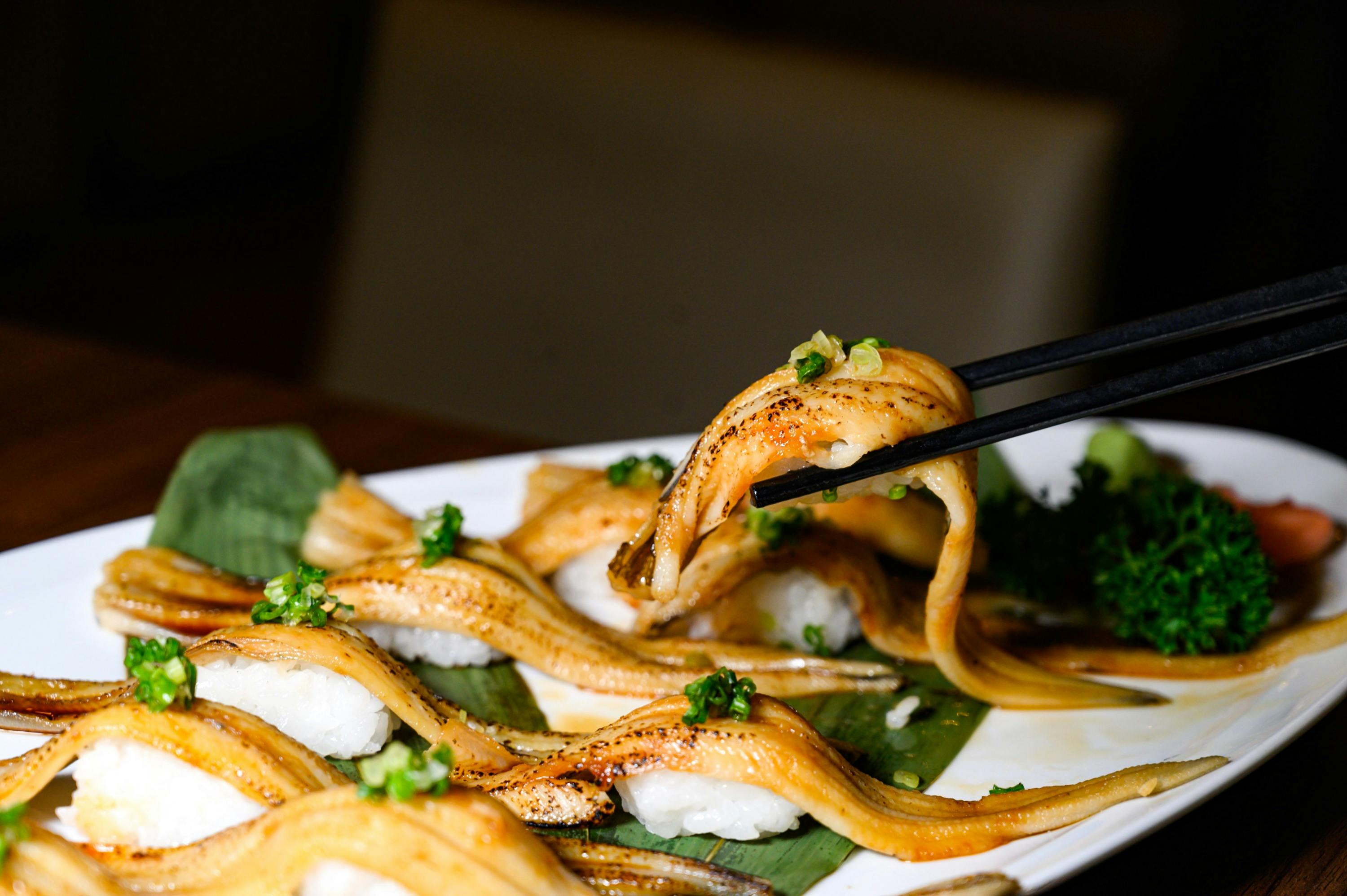 A plate of sushi topped with grilled eel and green garnish, with chopsticks lifting one piece.