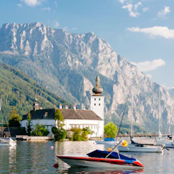 Båtar flyter på en sjö med en vit byggnad med ett torn på stranden. Berg reser sig i bakgrunden under en blå himmel.