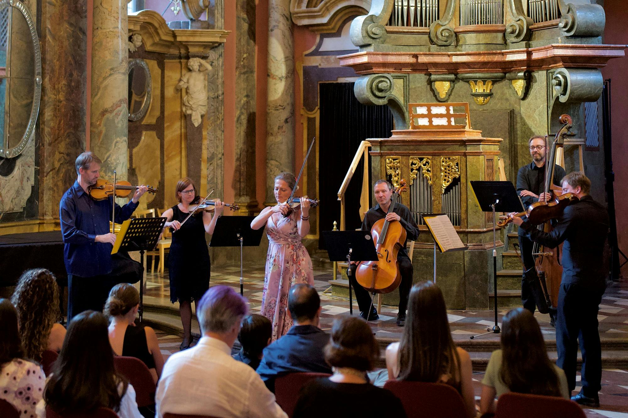 Musicians perform with string instruments in an ornate hall, while an audience sits and watches attentively.