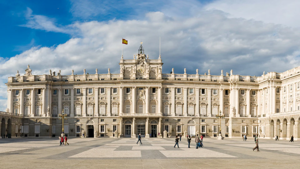 People walking in a large courtyard in front of an ornate, multi-story building with arches, columns, and a flag on top.