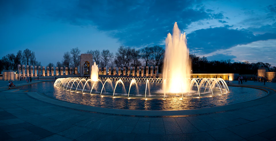Illuminated fountains at dusk with a backdrop of trees and an architectural structure with arches.