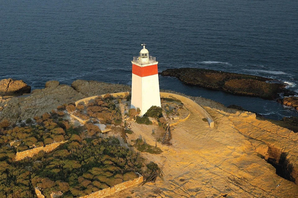 Aerial view of a white and red lighthouse on a rocky coastal area with surrounding vegetation, next to the ocean.
