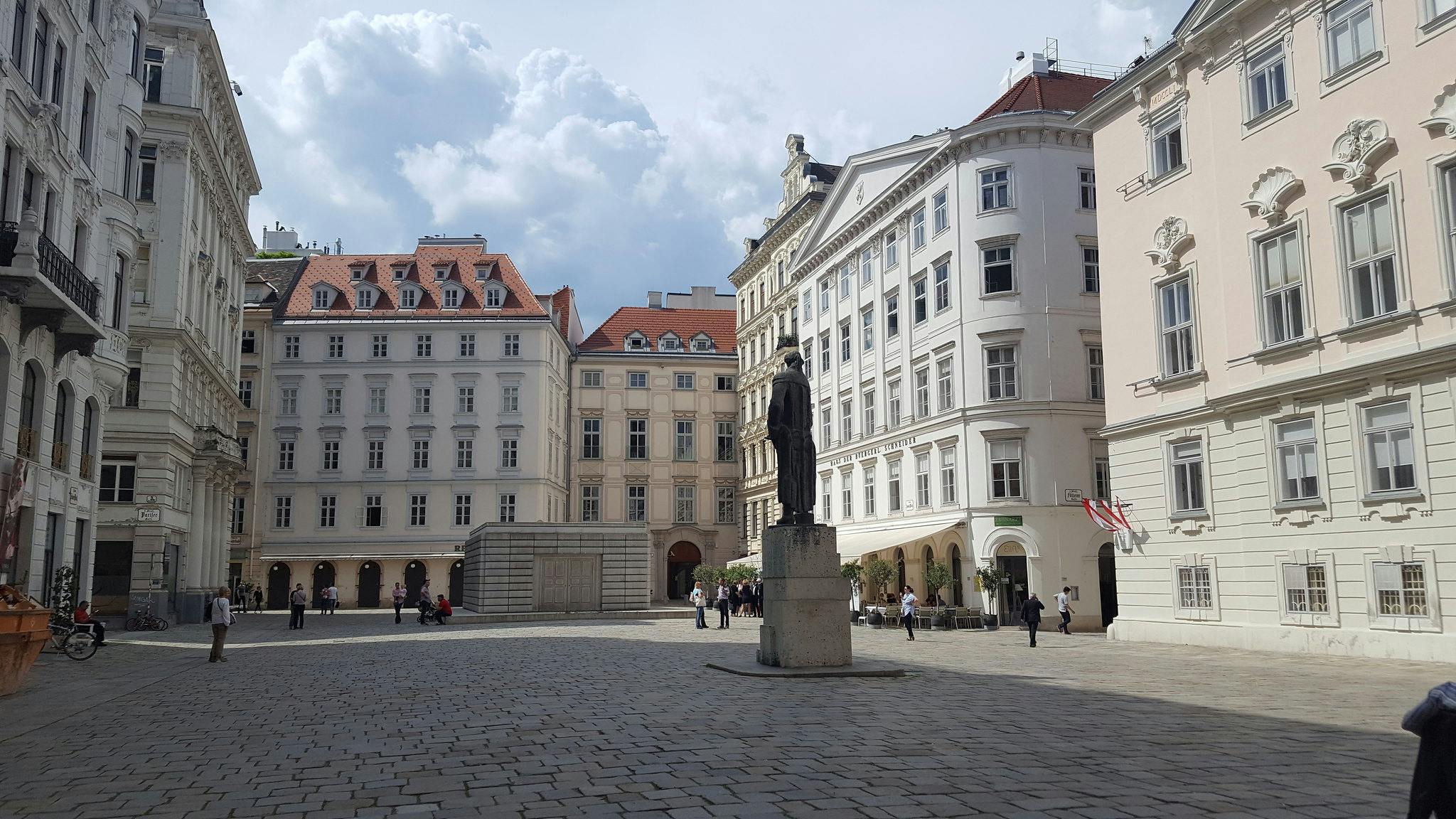 A European square with historic buildings, a statue in the center, and people walking and sitting on benches around the plaza.