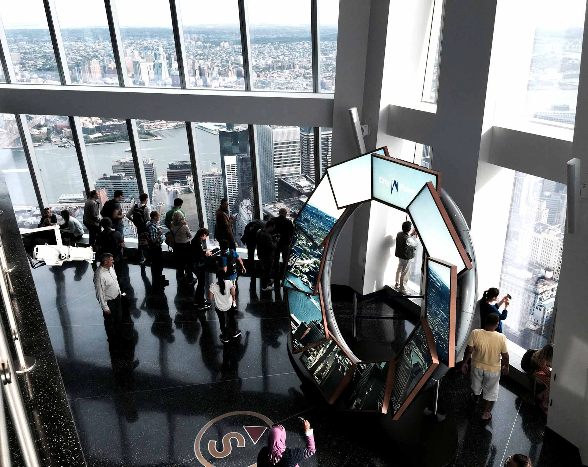People gather at an observation deck with large windows, providing a view of a cityscape and river. A multi-screen display stands nearby.