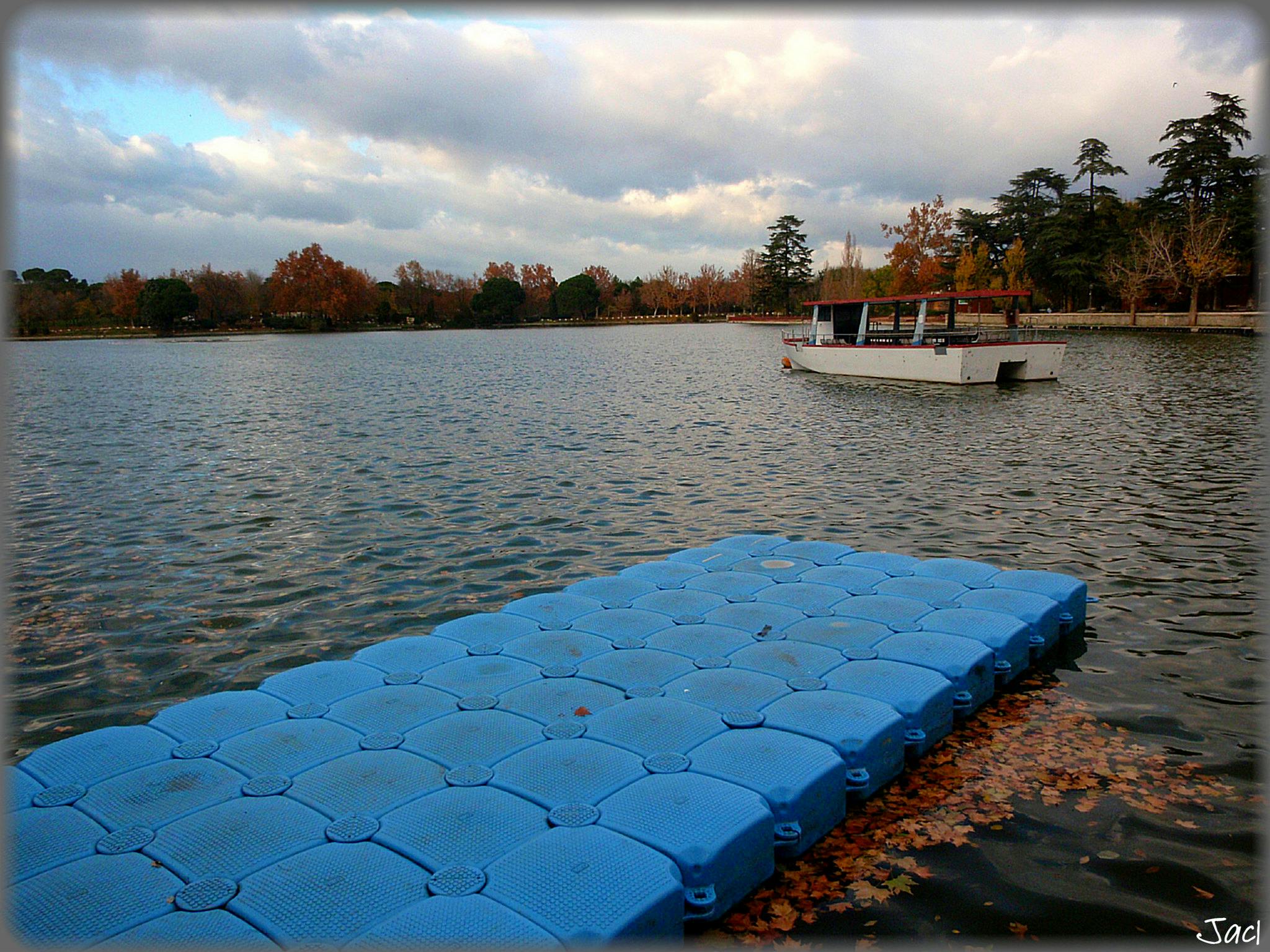 En lugn sjöscen med en flytande blå brygga och en tom vit båt under en molnig himmel, omgiven av höstträd.
