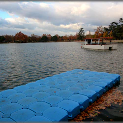 En lugn sjöscen med en flytande blå brygga och en tom vit båt under en molnig himmel, omgiven av höstträd.