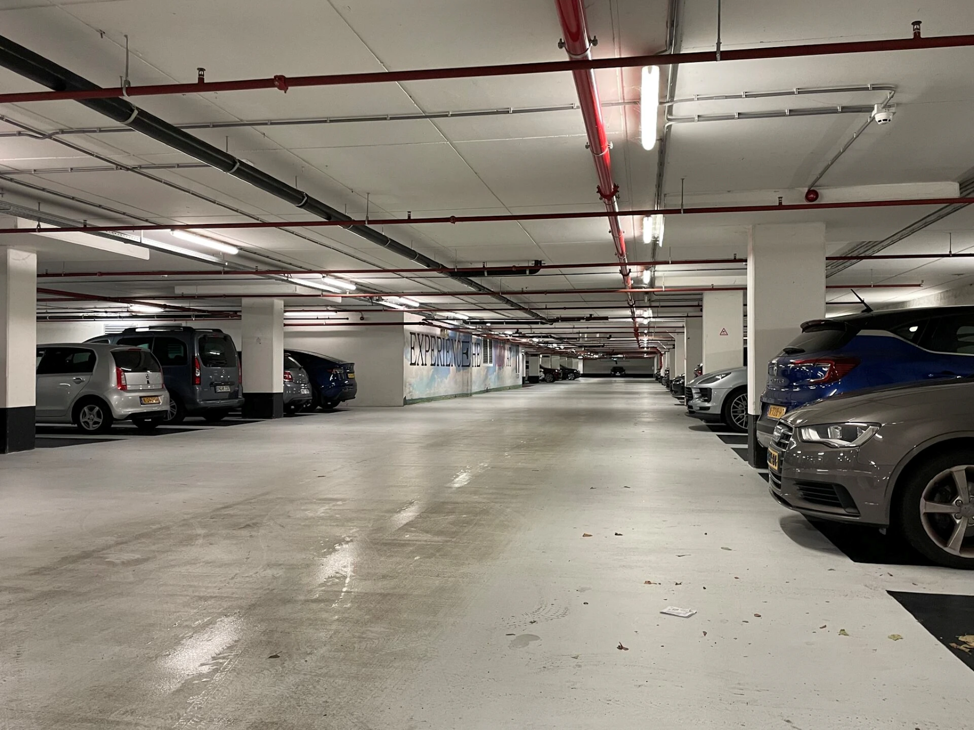 Underground parking garage with multiple parked cars, white walls, red pipes on the ceiling, and overhead fluorescent lighting.