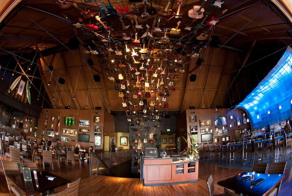 A restaurant interior with a wooden ceiling and numerous guitars hanging above, surrounded by tables and display cases.