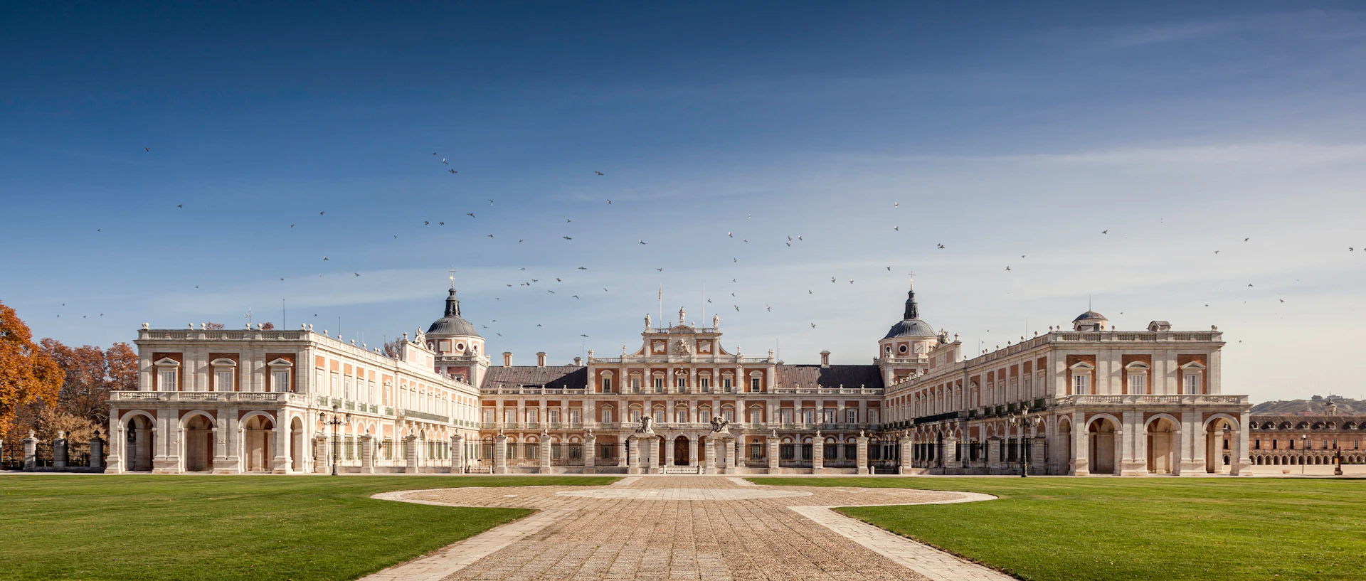 A grand palace with two domed towers, a central building, and a wide courtyard, under a clear sky with scattered birds.