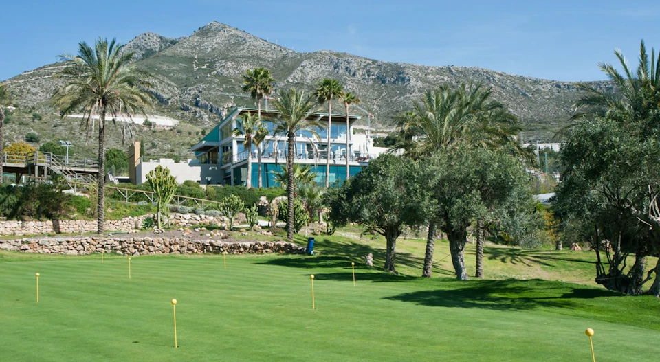 Modern building surrounded by palm trees and greenery, located in a mountainous area with a manicured lawn in the foreground.