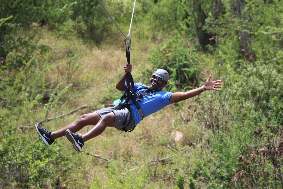 A person ziplining through a forested area, smiling and wearing a helmet, blue shirt, and harness.