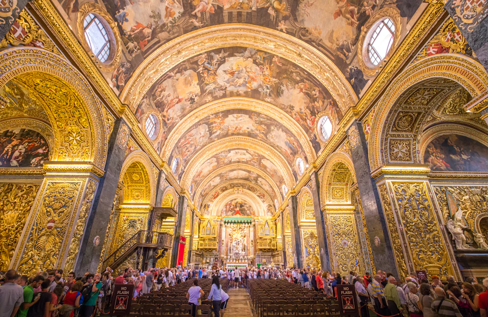 Opulent church interior with detailed frescoes on arched ceilings, ornate gold decorations, empty chairs, and people observing.