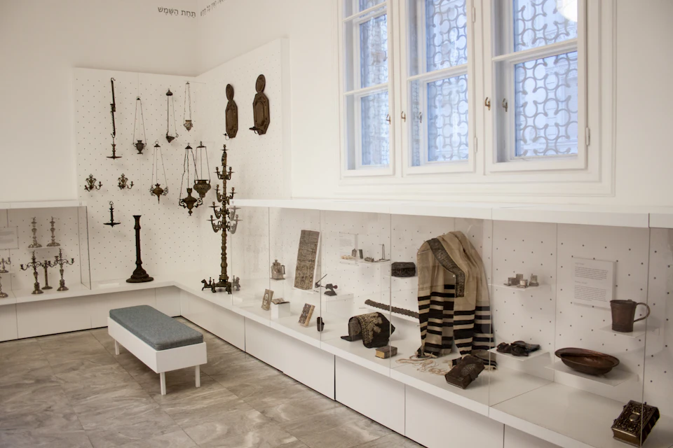 Museum display room with religious artifacts, including hanging candlesticks, prayer shawls, and books, beneath large windows.