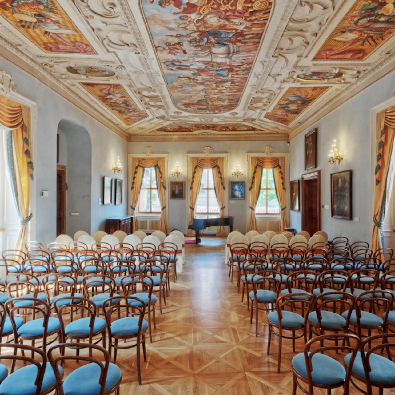 Elegant room with ornate ceiling frescoes, rows of wooden chairs with blue seats, large windows with draped curtains, and framed paintings.