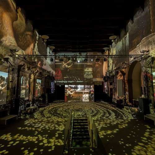Large, dark room with immersive digital projections of artwork on walls and floor, creating a vibrant and dynamic visual display.