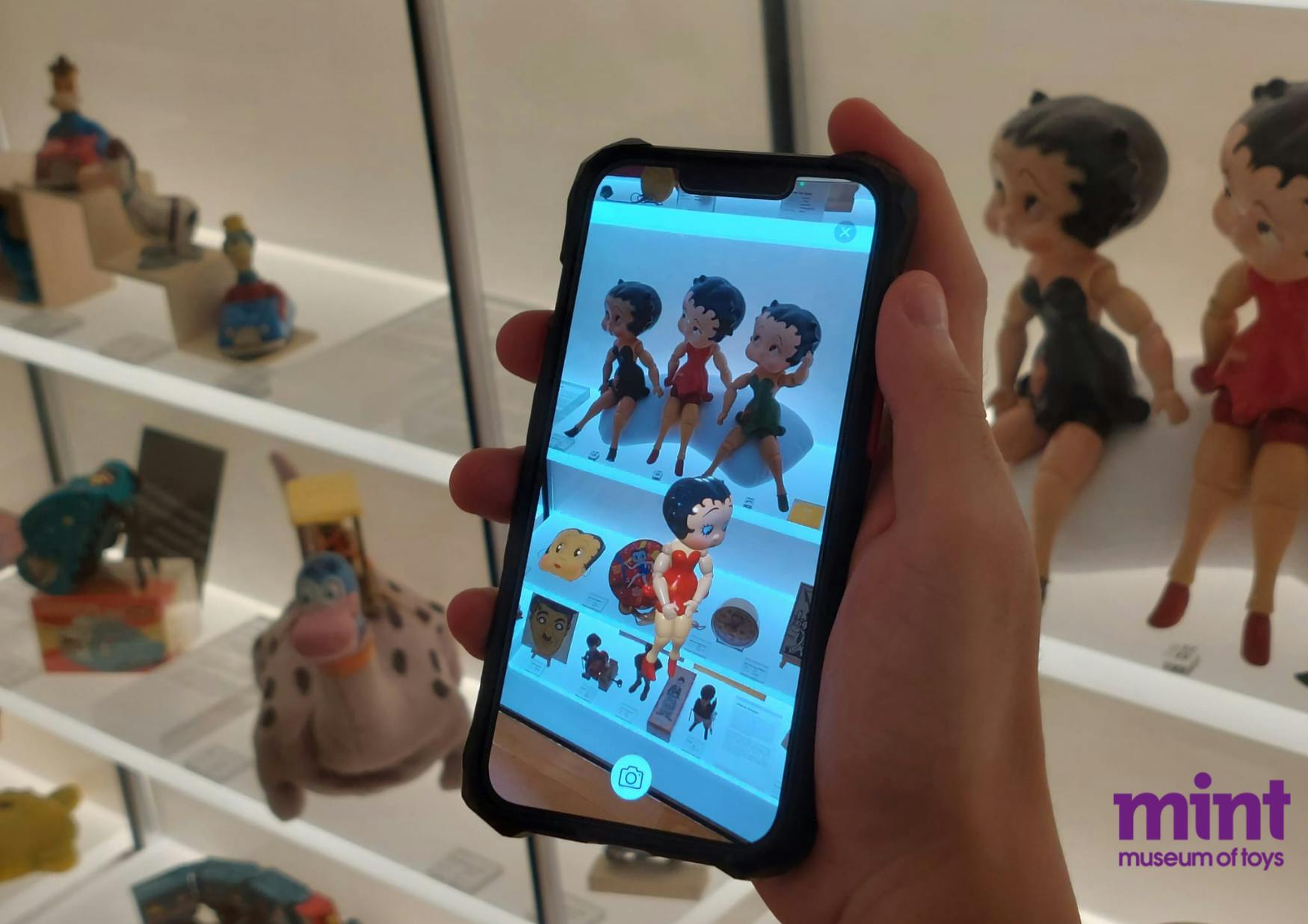 人形ディスプレイの前にベティ・ブープ人形の写真を表示しているスマートフォンを持った手があり、隅には「MINT MUSEUM OF TOYS」があります。