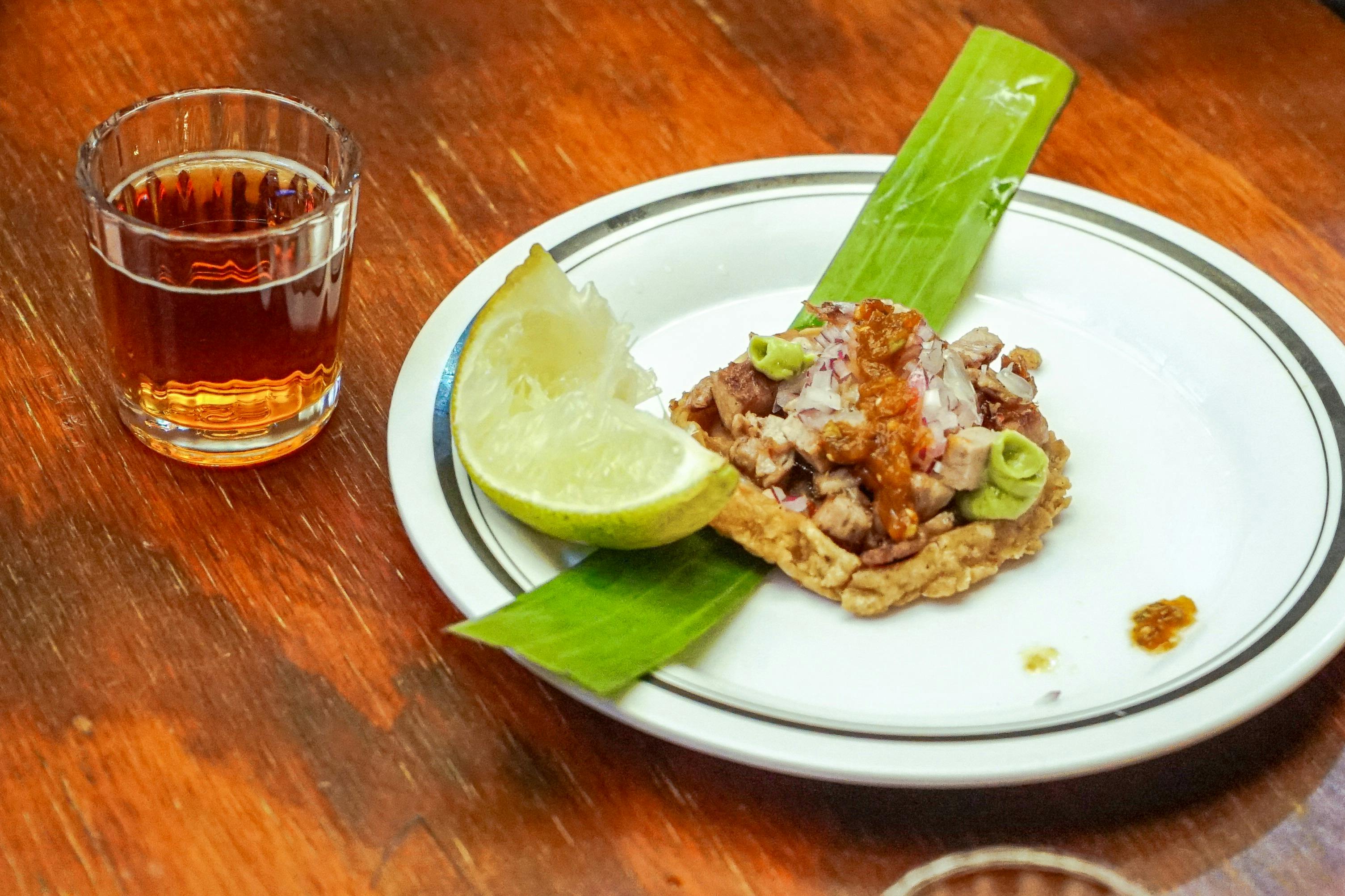 Un plato con un taco pequeño, adornado con lima y salsa, junto a un vaso de bebida sobre una mesa de madera.