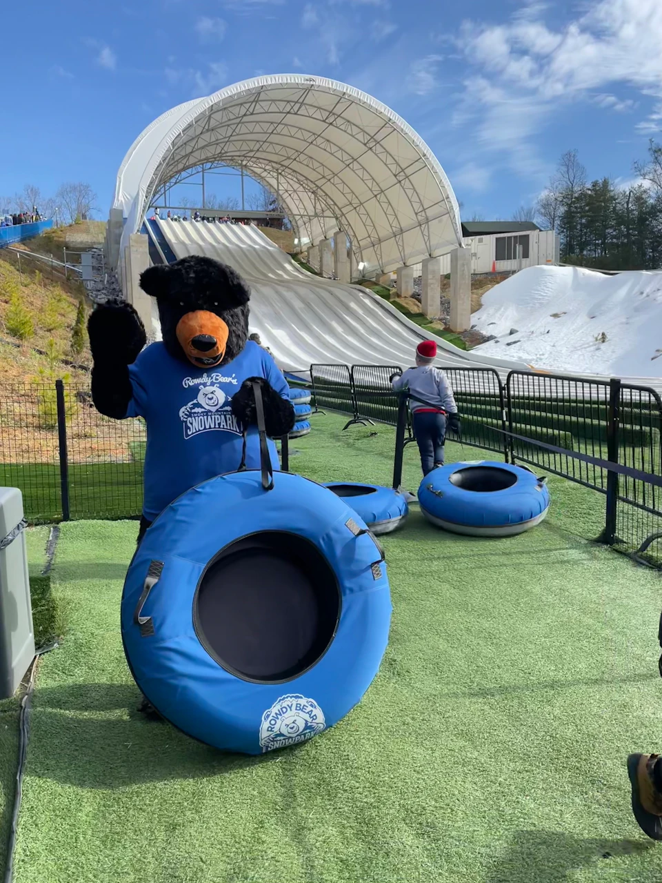 Bear mascot standing on grass with snowtubing slopes in the background. Another person is climbing up with a blue tube.