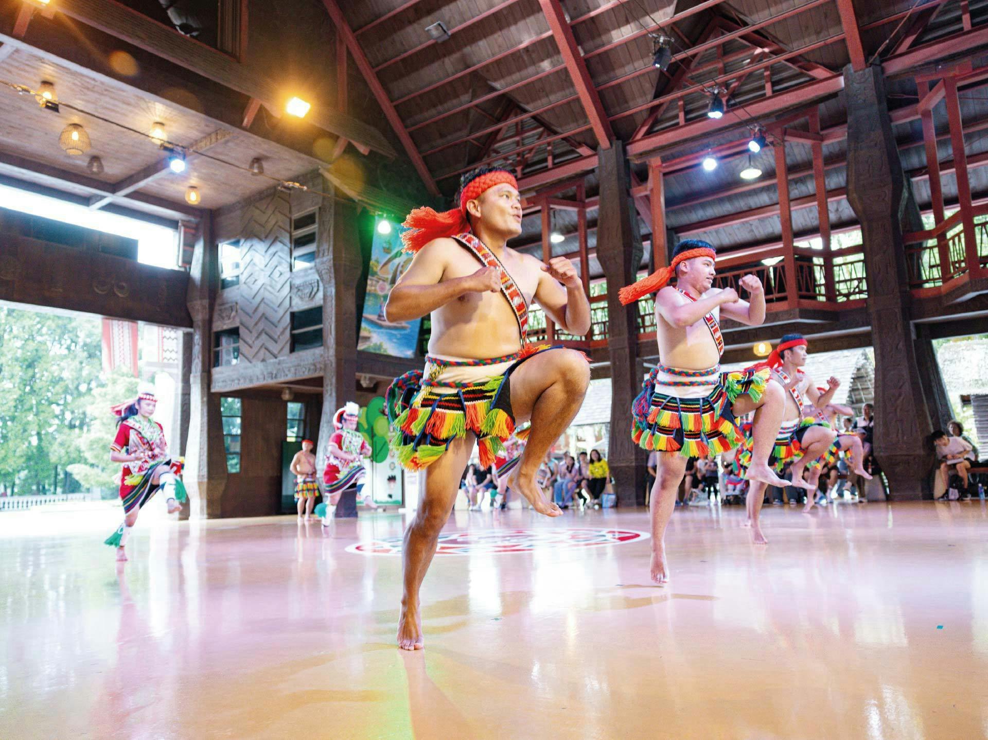 A group of performers in colorful traditional costumes dancing energetically inside a wooden building with bright lighting.