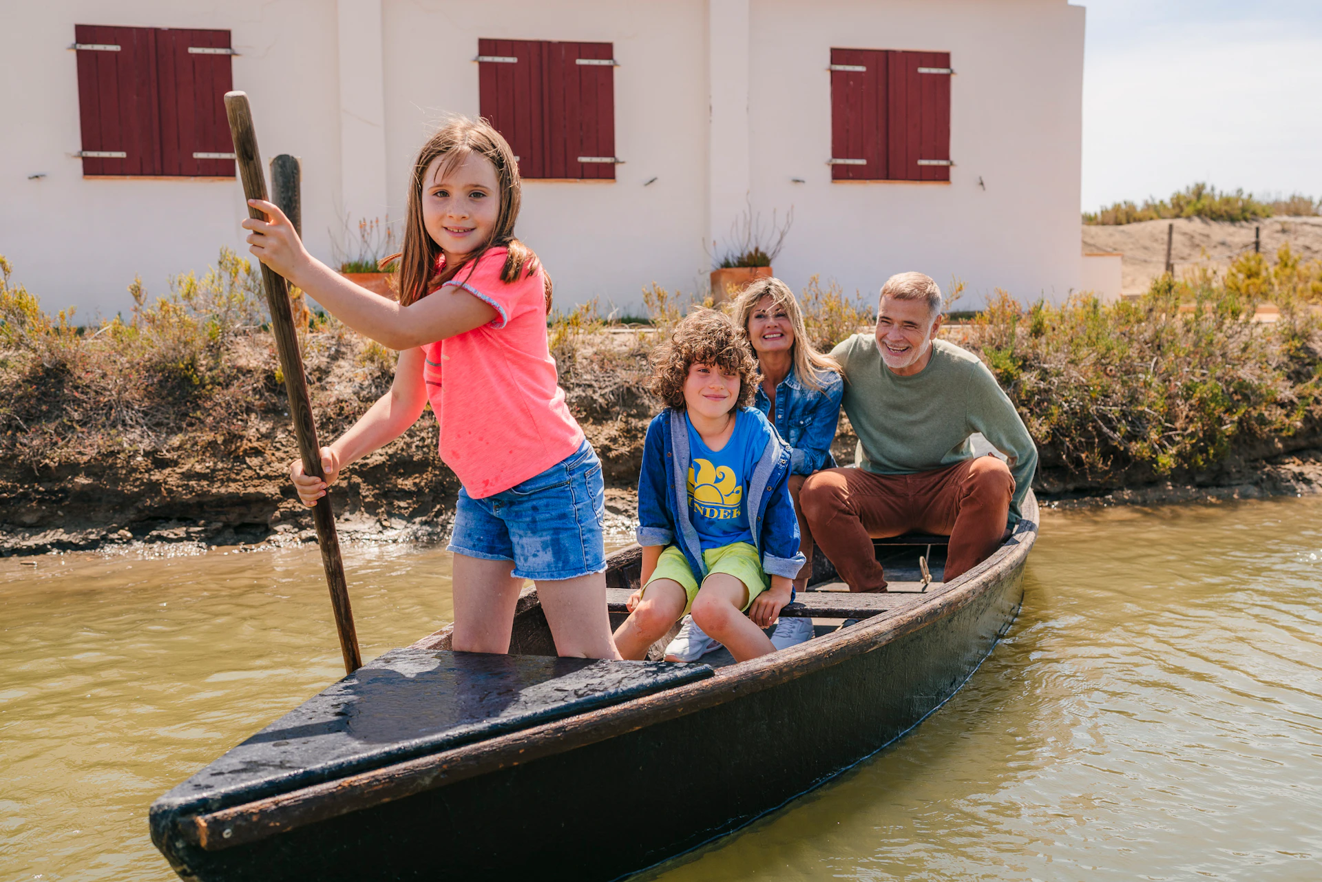 A family of four, two adults and two children, are enjoying a boat ride on a narrow river near a white building with red shutters.