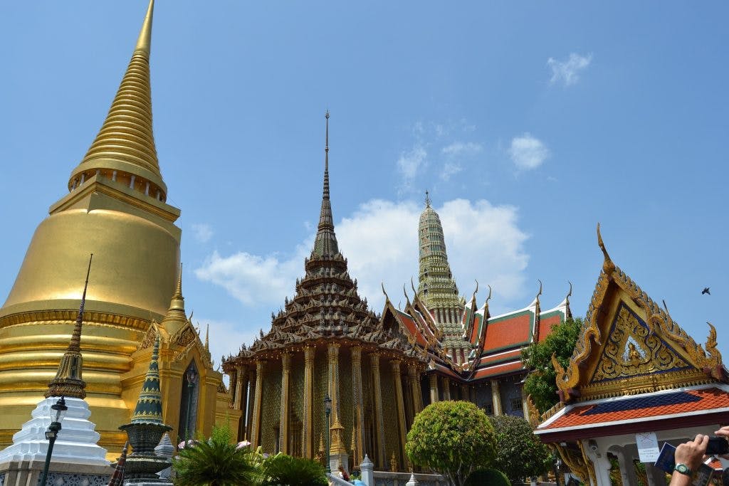 A temple complex with ornate spires, golden stupa, red-roofed buildings, and green foliage under a clear blue sky.