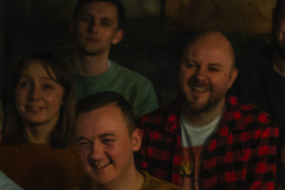 A group of people watch a person covered in fake blood and lying on a table in a rustic, dimly lit room.