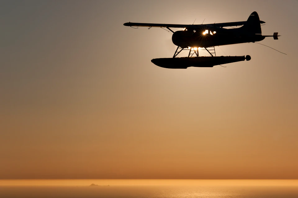 Silhouette of a seaplane flying over calm water at sunrise or sunset, with light shining through the plane's windows.