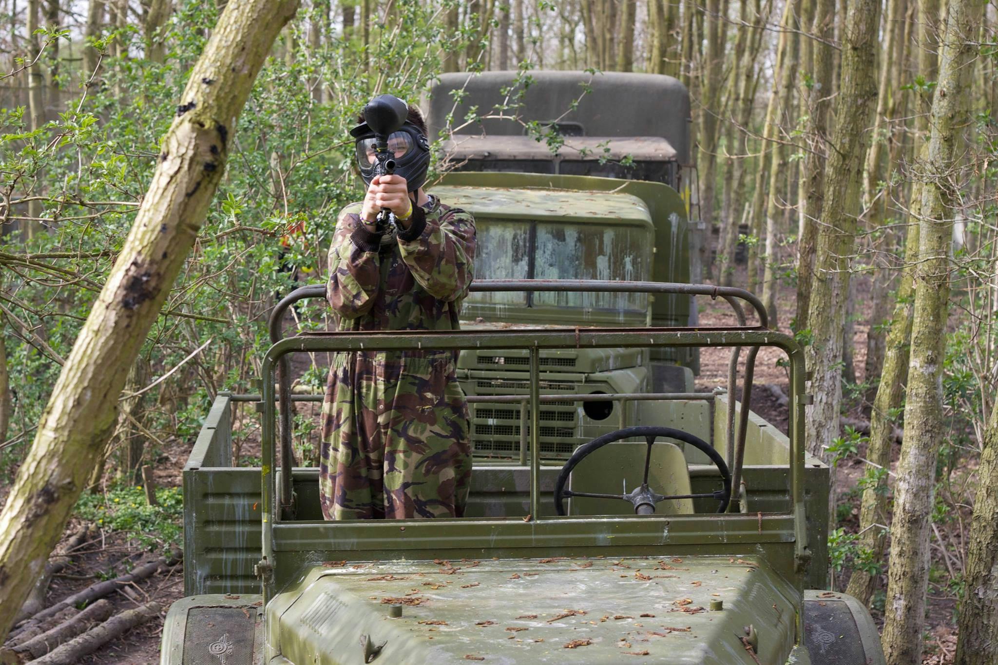 Ένα άτομο με μάσκα καμουφλάζ και paintball που στοχεύει ένα όπλο paintball στέκεται μπροστά από ένα φορτηγό στρατιωτικού τύπου σε μια δασώδη περιοχή.
