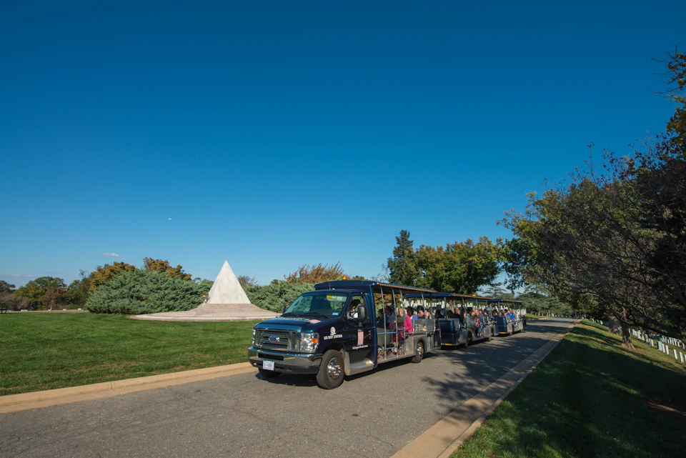 A tour tram filled with people drives along a paved road in a park, with a white pyramid structure and trees in the background.
